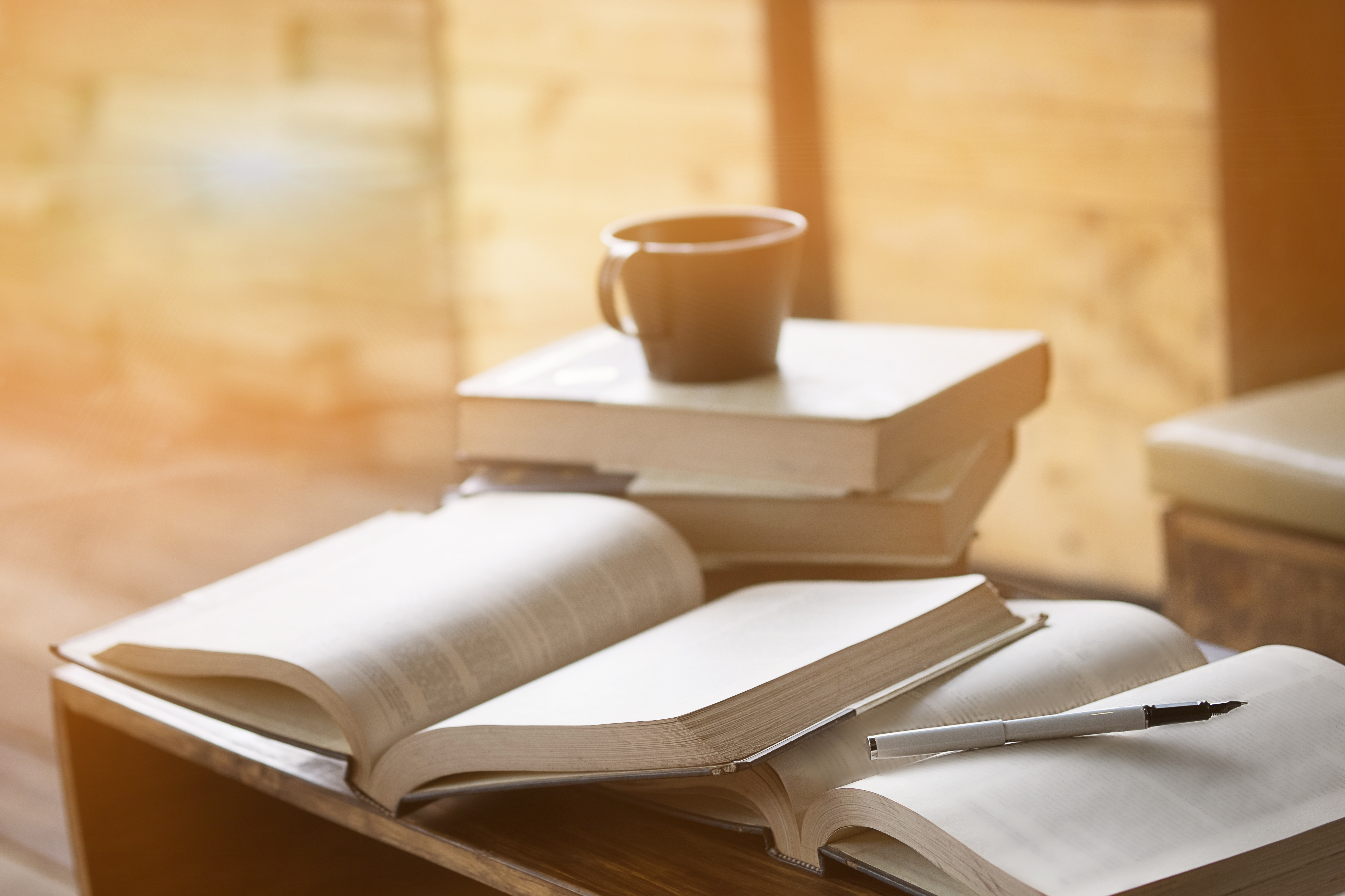 A stack of open and closed books on a wooden table, with a coffee cup and a pen placed nearby, bathed in warm ambient light.