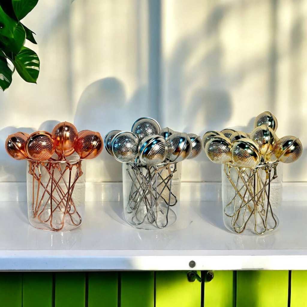 A collection of metal strainers in various finishes, including copper, silver, and gold, arranged in clear containers on a white surface.