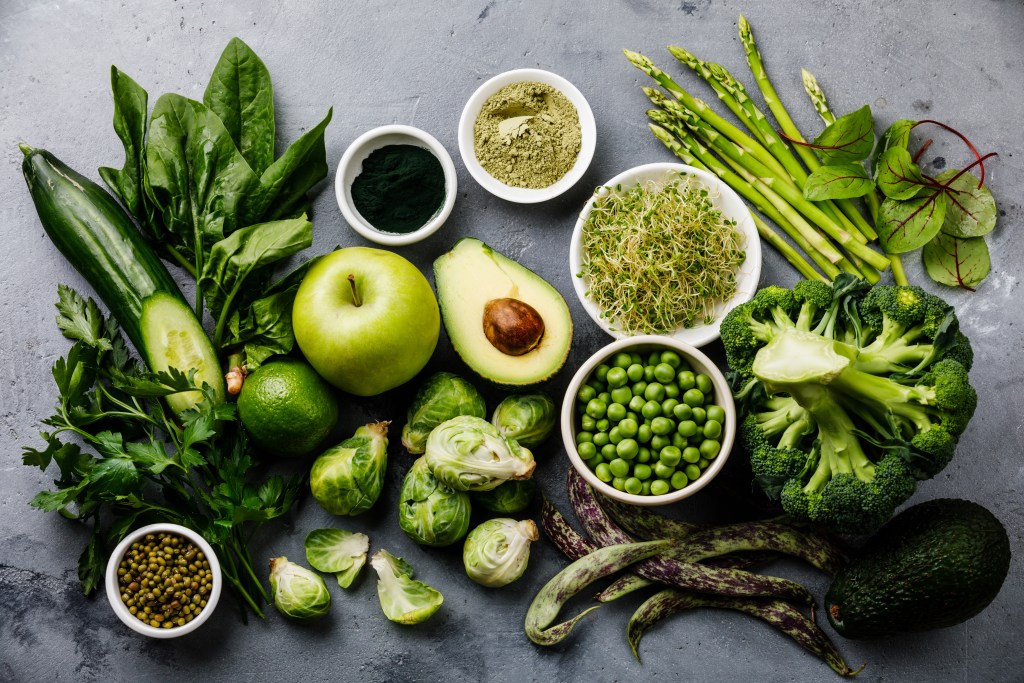 An array of various fresh green vegetables, including cucumbers, spinach, broccoli, Brussels sprouts, and green apples, arranged artistically on a textured surface.
